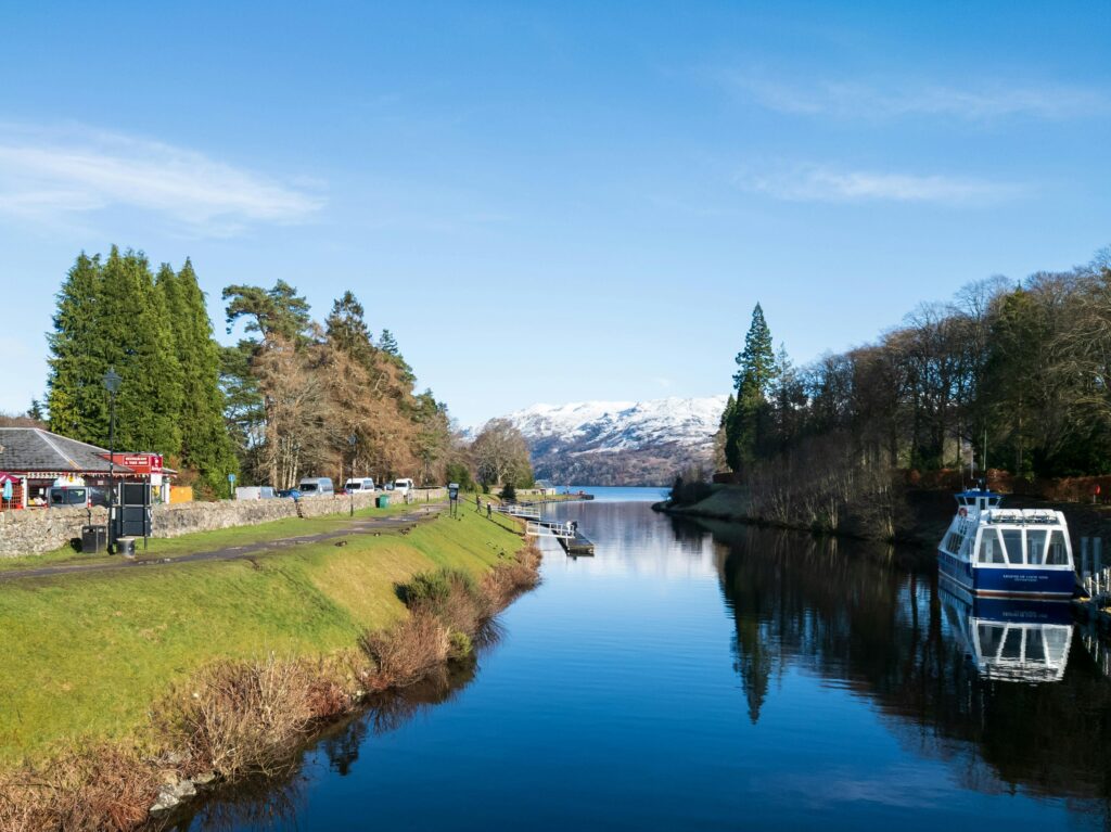 A peaceful view of the Caledonian Canal with a boat, surrounded by greenery and snowy mountains in Fort Augustus, Scotland on a private tour of Scotland from Edinburgh on a tailor made itinerary