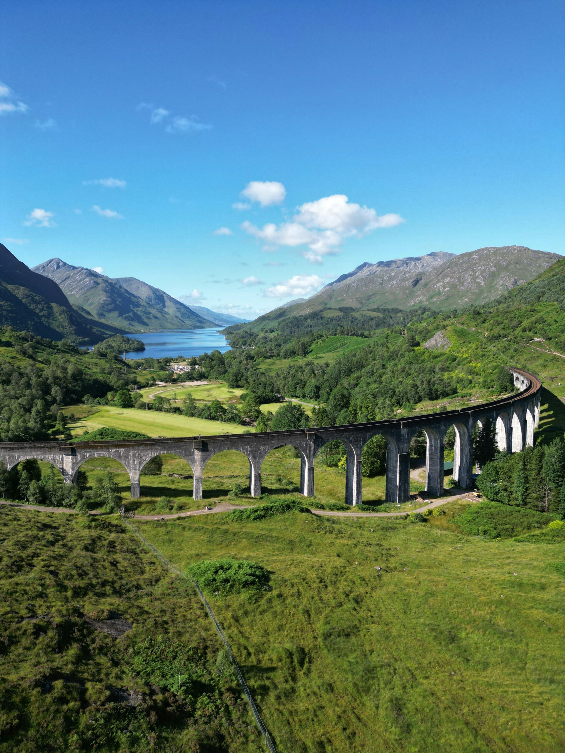 A breathtaking view of the Glenfinnan Viaduct surrounded by lush green mountains and a clear blue sky. A great private tour for Harry Potter fans, to see the hogwarts express or Jacobite steam train