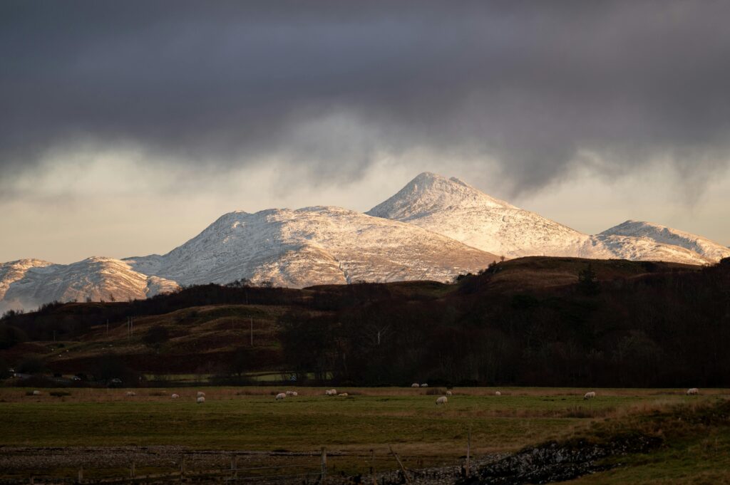 Breathtaking view of snow-capped mountains with fields, captured under a dramatic cloudy sky. Ben Nevis View in the Scottish Highlands on a private tour to the Scottish Highlands from Edinburgh
