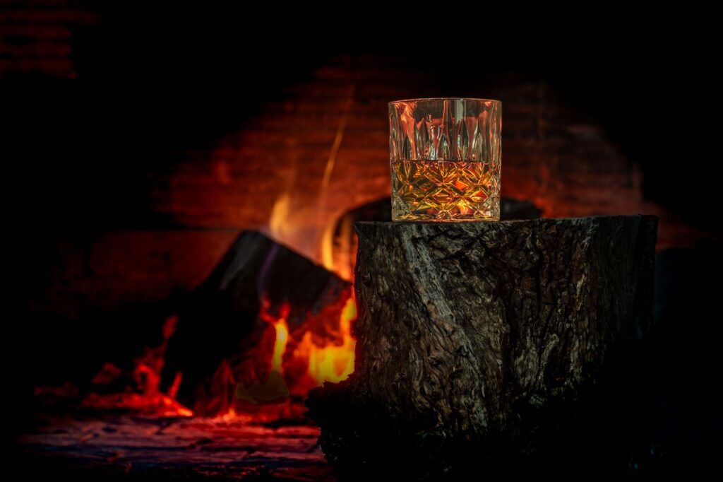 Close-up of whisky glass on a tree trunk by a warm fireplace, creating a cozy atmosphere.